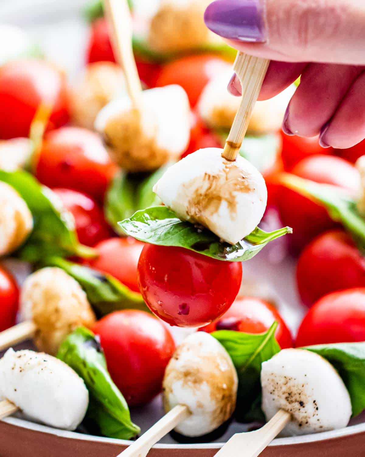 and hand holding a caprese salad bite on a pick over a plate full of them